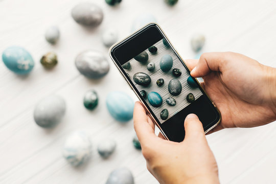 Hands holding phone and taking photo of stylish easter eggs on rustic white wooden background. Modern easter  eggs painted with natural dye in grey marble color. Happy Easter