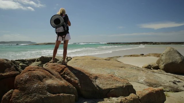 Blonde backpacker woman photographer holding her camera at William Bay National Park, Denmark, Western Australia. Tropical destination Madfish Beach surrounded by rocks. Australia summer holidays.