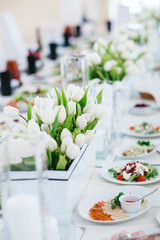 Dinner table serving. White tulips stand in flowerpots on white table served with delicious food