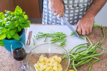 top view of man's hands cutting with a steel knife some fresh green beams to eat something healthy and perfect for diet - red wine and basilico and potatoes on the table ready to be cooked at home