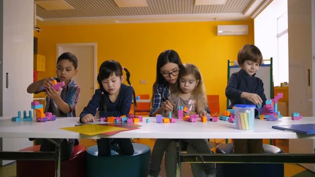 Group of diverse multinational preschool children playing with colorful construction toy blocks, expressing their creativity and ideas in classroom under the guidance of young kindergarten teacher.