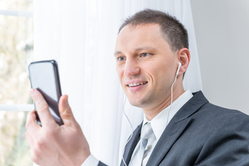 Closeup of young, happy man, male businessman in business suit, tie, standing by window in home, house room, white curtains, with earbuds headphones, holding phone, talking, video conferencing