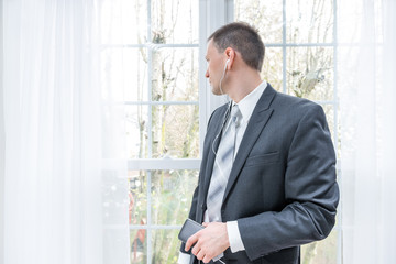 Closeup of young, happy smiling man, businessman, business suit, tie, standing by window in home, house room, white curtains, with earbuds headphones, talking, music listening, holding phone in hand