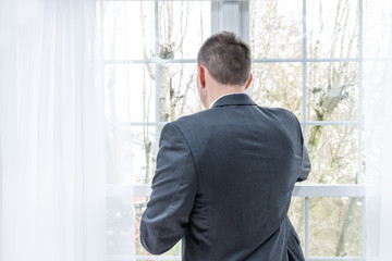 Closeup back of young man, businessman, business suit, tie, standing by window in home, house room, white curtains, with earbuds headphones, talking, listening to music on phone