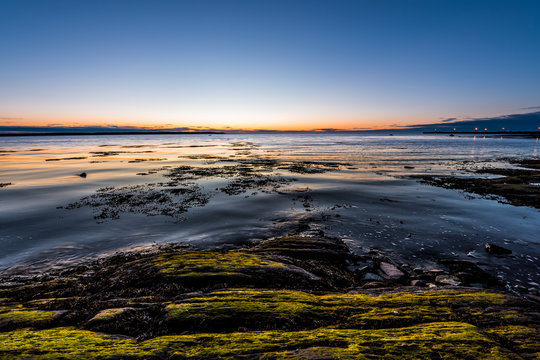 Twilight, dusk in Rimouski, Quebec, Saint Lawrence river, Gaspesie, Canada with rocks, boulders, rocky beach, turquoise water, sun reflection seaweed blue sky