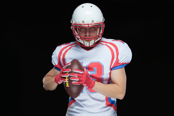 American football player wearing helmet posing with ball on black background