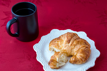 Fresh croissant on a white stoneware plate on red tablecloth with black coffee up, top view