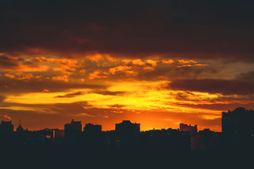 Cityscape with vivid fiery dawn. Amazing warm dramatic cloudy sky above dark silhouettes of city buildings. Orange sunlight. Atmospheric background of sunrise in overcast weather. Copy space.