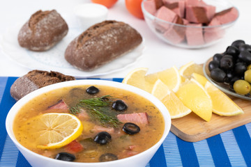 Traditional Russian soup Solyanka cooked with meat, sausages, salted cucumbers on wite bowl. Black bread, lemon, olived and tomatoes on color blue background, homemade food.