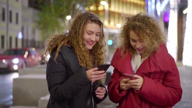 Two beautiful women laughing looking at funny stuff on smartphones outdoor in stylish clothes.