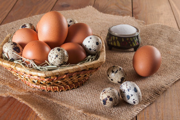 Some brown chicken eggs and several small motley quail eggs on straw in wicker basket and three quail eggs, one chicken egg and salt on sackcloth. All this on wooden table.