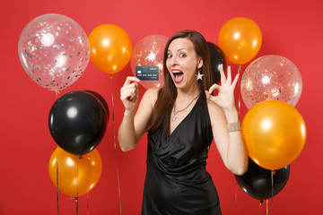 Excited young woman in little black dress celebrating, holding credit card, showing OK sign on red...