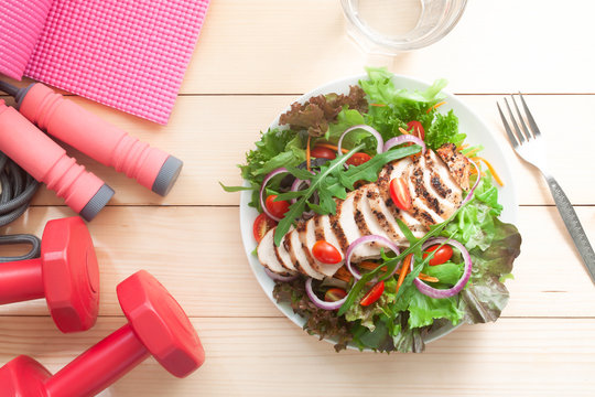 Fresh salad with chicken and fitness exercise equipments on wooden table