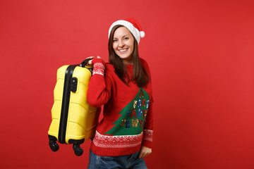 Smiling young Santa girl in knitted sweater, Christmas hat holding yellow suitcase isolated on...