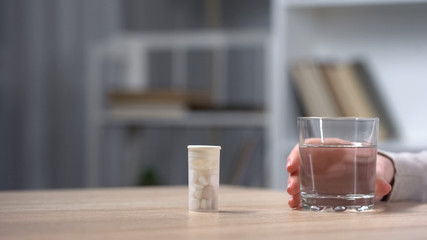 Medicine tube standing on table, female hand holding glass of water, healthcare