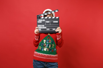 Shocked young Santa girl hiding, covering face with classic black film making clapperboard isolated...