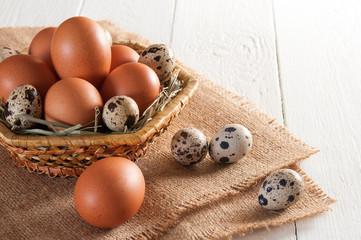 Quail and chicken eggs on straw in a basket, near three quail eggs and one chicken egg on sackcloth. On white wooden table