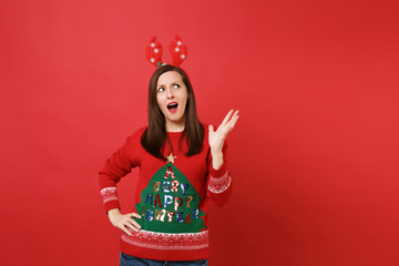 Outraged young Santa girl in fun decorative deer horns on head looking up rising hand isolated on bright red wall background. Happy New Year 2019 celebration holiday party concept. Mock up copy space.