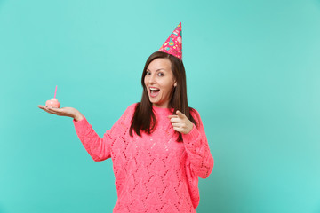 Cheerful young woman in knitted pink sweater, birthday hat pointing index finger on camera hold in...