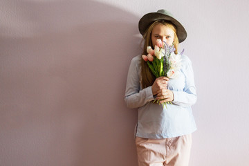 A young beautiful girl with blond dissolved long hair, a felt hat on her head, keeps spring flowers...