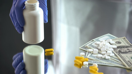 Hand holding pills jar, dropping tablets on dollar, expensive medical treatment