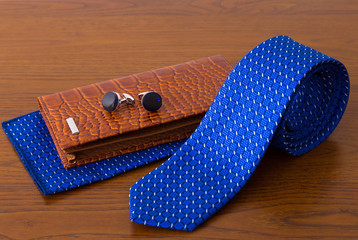 Brown leather wallet, couple of metal cufflinks, blue necktie with pocket square on wooden table....