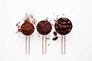 Rose gold measuring cups of cocoa beans, cacao nips and cocoa powder on a white background, flat...