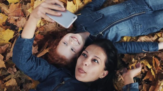 A beautiful mother and her cute daughter lie on a yellow leaf in the autumn park and making a selfie on smartphone. Happy family is having fun. Top view, slow motion