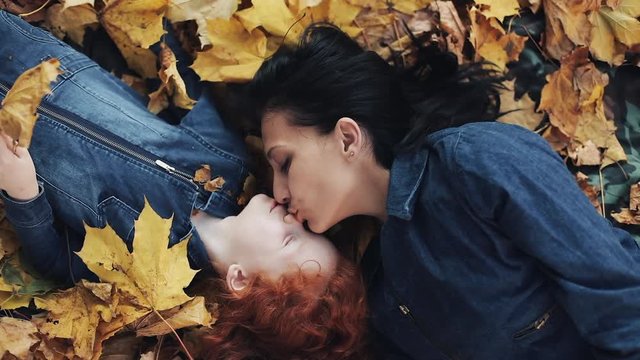 A beautiful mother and her cute daughter lie on a yellow leaf in the autumn park. Mom kisses daughter. Happy family is having fun. Top view, slow motion