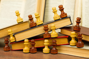 Black and white chess pawns arranged chaotically on stack of vintage books on wooden table 