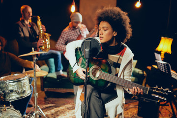 Mixed race woman singing and playing guitar while sitting on chair with legs crossed. In background...