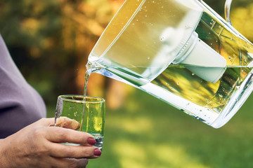 Pouring filtered water from a water filtration jug into a glass which female hand holding in summer...