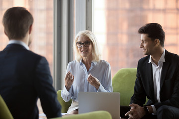 Business people sitting on comfortable couch in contemporary office near panoramic window. Middle aged company ceo and middle eastern ethnicity colleague talking with client discussing working details