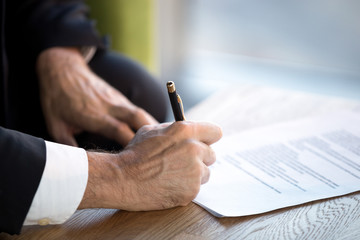 Confident businessman sitting in office desk holding pen puts his signature on official document, close up of male hands and table. Beneficial cooperation, important decision, financial deal concept