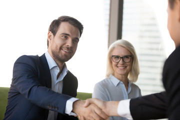 Business people greeting each other at meeting, focus on handsome young confident businessman welcoming client. Aged female in eyeglasses, team leader and company partner handshaking in modern office
