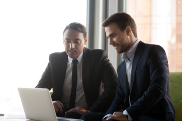Aged and young businessman at meeting in modern office. Colleagues or client and executive manager sitting on couch looking at laptop screen working together analyzing sales statistics using program
