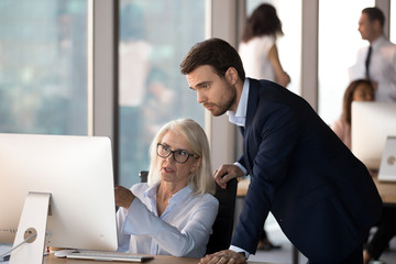 Obraz premium Business people using computer, millennial male employee in formal suit helping explaining to middle aged mature female colleague learn understand corporate program. Mentoring and assistance concept