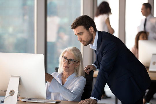 Business people using computer, millennial male employee in formal suit helping explaining to middle aged mature female colleague learn understand corporate program. Mentoring and assistance concept
