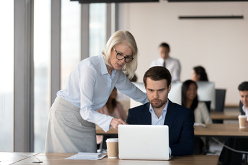 Obraz premium Middle aged female executive manager and male colleague working together in coworking space, businesswoman pointing on computer screen analyzing explaining business project results and discussing data