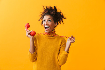 Portrait of happy african american woman with afro hairstyle screaming while holding red handset, isolated over yellow background