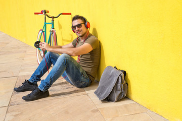 Fototapeta na wymiar Young man wearing sunglasses relaxing on the floor and listening music. Sitting against a yellow wall with his computer