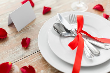 valentines day, table setting and romantic dinner concept - close up of plates with cutlery, red rose petals and blank place card
