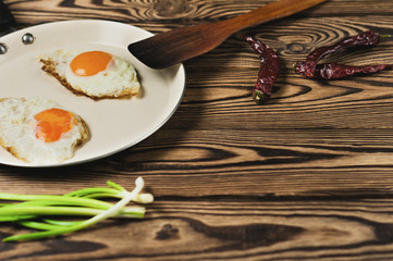 Two fried eggs in metal beige pan near dirty spatula and green fresh onion and dried red hot pepper on old wooden brown table. Copy space