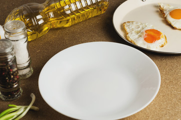 Two fried eggs in metal beige pan near white empty ceramic plate and bottle with oil and spices
