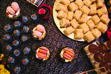 Candy bar table set. Cookies, eclairs, macaroons, fruits, cream and layered cakes. Black, golden and red colours.