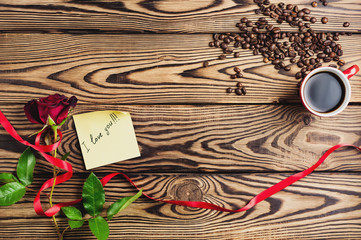 Inscription I love you on paper beside red rose with ribbon and cup of coffee near coffee beans on old wooden table. Top view with copy space. Saint Valentine's Day concept