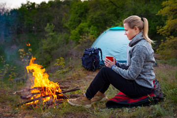 Woman traveler resting by the fire on the background of a tent on his footpath through the mountains and the forest