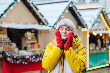 Outdoor close up portrait of young happy smiling girl holding by cheeks in knitted hat, red mittens, yellow jacket having fun, enjoy snowfall in street of european city. Winter mood, holidays concept.