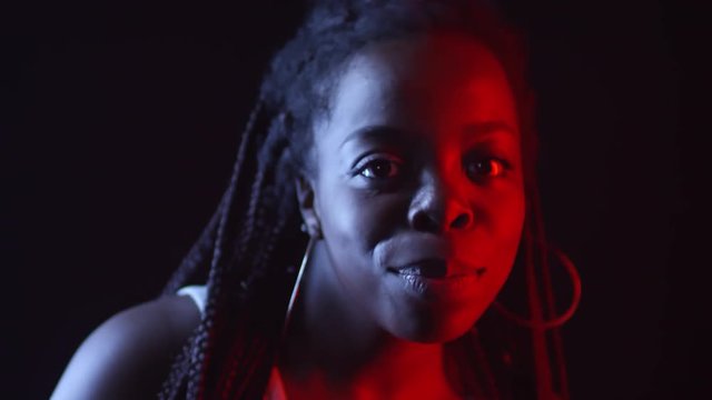 Low-key portrait shot with red and blue lighting: authentic black woman with braids and hoops earrings dancing isolated on dark background and smiling for camera