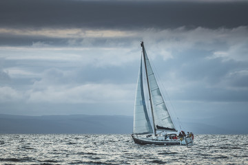 Lone touristic yacht sailing in the sea. Dark cloudy sky before the rain. Northern Ireland. Amazing marine scenery the small ship among the calm water. Ocean scape in grey blue tints.